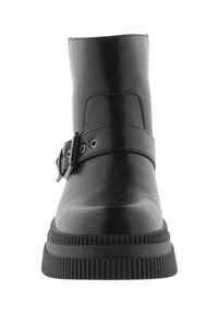 Bota de cuero negro hasta el tobillo con un acabado suave, suela de goma plana y correa ajustable con herrajes metálicos y recortes circulares.