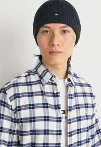 Schwarze gerippte Mütze mit einem Logo, gestreifte Flanellhemd in Weiß und Marineblau und ein weißes T-Shirt mit Text darunter, mit sichtbaren Knöpfen.