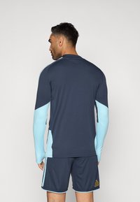 Ναυτικό αθλητικό μπλουζάκι με ανοιχτό μπλε μανίκια και πλαϊνές πάνελ. Διαθέτει ραμμένο γιακά και λογότυπο Adidas στα σορτς. Λείος, εφαρμοστός υφή.