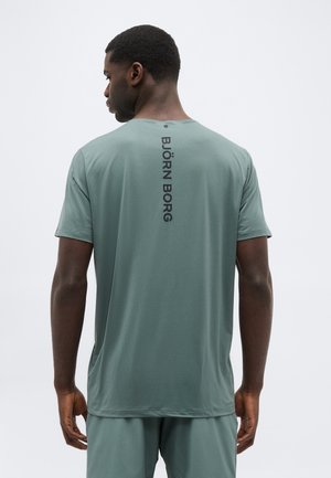 Mees, kes kannab helerohelist lühikese varrukaga särki ja lühikesi pükse, näitab tagakülge, kus särgil on vertikaalselt trükitud tekst "BJÖRN BORG" ühevärvilise tausta ees.