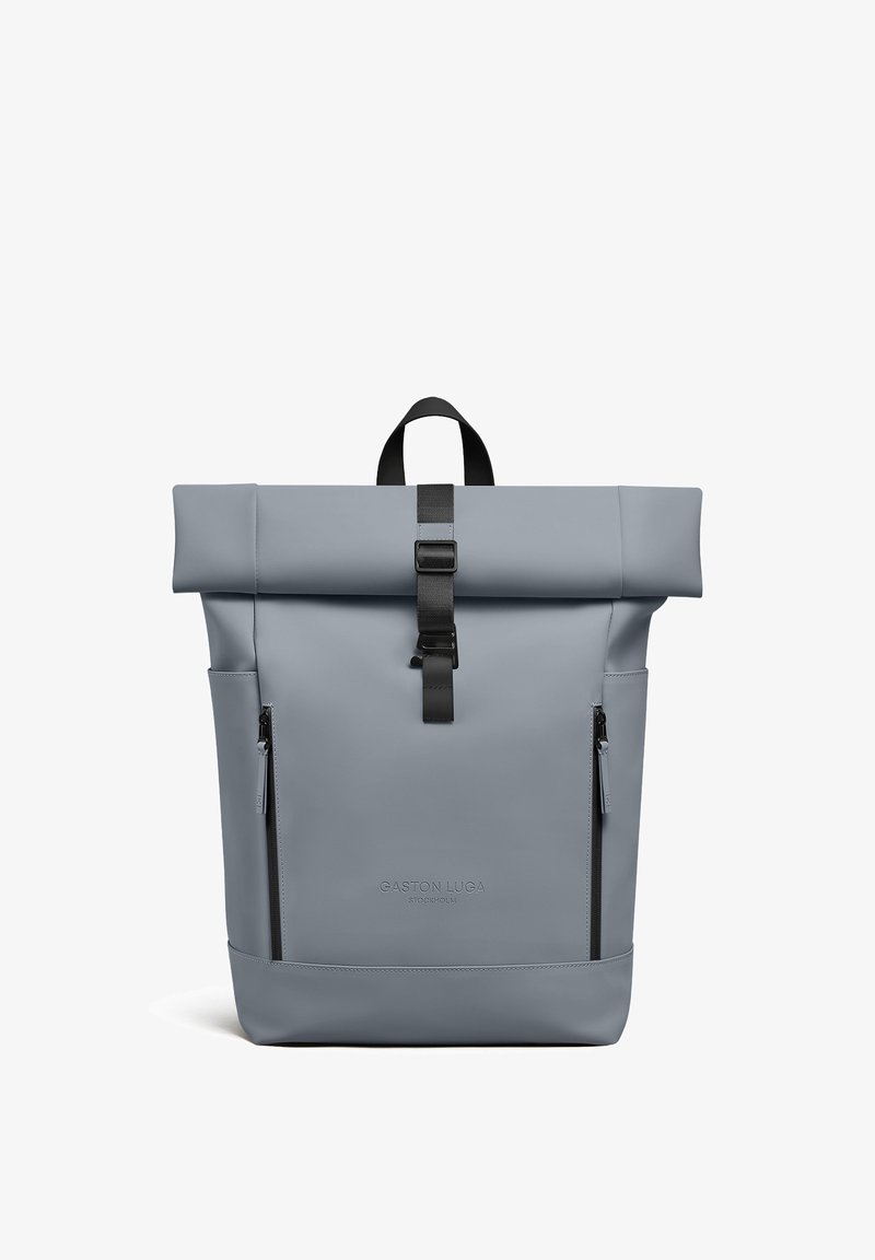 Sac à dos gris clair à rabat enroulé avec boucle noire, deux poches zippées verticales et logo Gaston Luga Stockholm sur le devant.