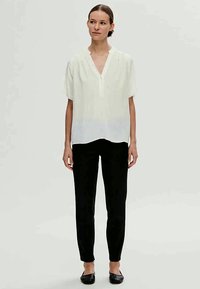 Blusa branca de mangas curtas com decote em V e tecido ligeiramente transparente, combinada com calças pretas de corte justo e sapatos planos pretos.