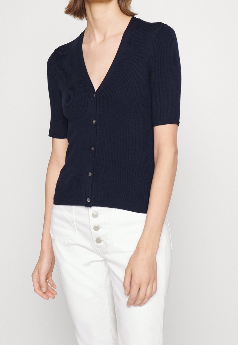 Marinblå ribbstickad kofta med djup V-ringning och korta ärmar. Har knäppning fram och subtil textur. Kombineras med vita byxor med hög midja.