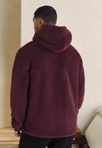 Bordeauxkleurige hoodie fleece jack met een zachte, gestructureerde oppervlakte, met een relaxed fit en geribbelde manchetten, gezien van de achterkant.