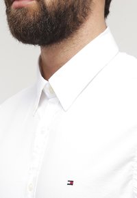 Witte overhemd met knopen en een klassieke kraag, met een klein geborduurd logo in rood en marineblauw op de borst. Glad stoftextuur.