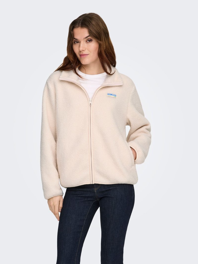 Femme portant une veste polaire beige clair zippée sur une chemise blanche et un jean bleu foncé, debout avec une main dans sa poche.