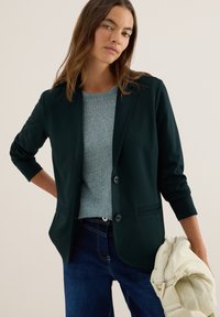 Donker groene blazer met een enkelvoudig ontwerp, twee knopen en een slim fit, gecombineerd met een lichtgrijze gestructureerde trui en denim spijkerbroek.
