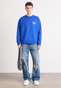 Sweatshirt azul com decote redondo e logo à frente, combinado com jeans claros rasgados e sapatilhas brancas, segurando uma mala cinza de ombro.