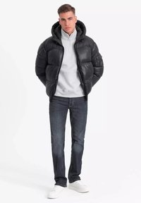 Zwarte pufferjack met een capuchon, glanzende textuur, rits sluiting, gedragen over een lichtgrijze sweatshirt, gecombineerd met donkere jeans en witte sneakers.