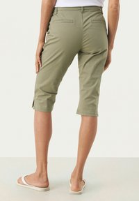 Pantaloni capri de lungime până la genunchi, de culoare verde măslin, dintr-un material moale, cu buzunare pe spate și detalii cu slit lateral, asortați cu sandale albe.