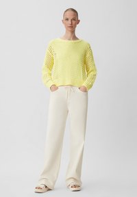 Limonengelben Strickpullover mit offenem Webdesign. Kombiniert mit cremefarbenen weiten Hosen; das Modell steht mit den Händen in den Taschen und trägt leichte Schuhe.