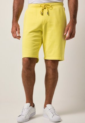 Mann in gelben Shorts mit Kordelzug und weißen Turnschuhen steht vor einem schlichten hellen Hintergrund.