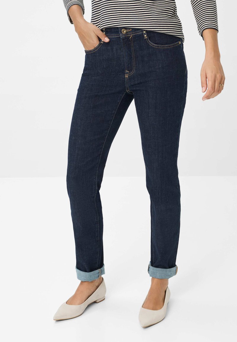 Jean en denim bleu foncé avec une coupe slim, cinq poches, des poignets retroussés et des coutures contrastées. Associé à des ballerines beige clair.