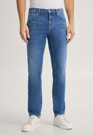 Man standing wearing blue straight-leg jeans, white sneakers, and a white shirt, shown from shoulders to feet against a plain background.