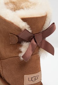 Ruskea mokkanahkasaapas, jossa on lampaanvillavuori, koristeellinen ruskea nauhakaarre ja UGG-logo-tarran sivussa.