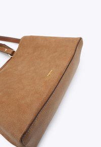 Sac à main en suede marron et cuir avec bandoulière réglable et petit logo doré sur le devant, sur fond blanc.