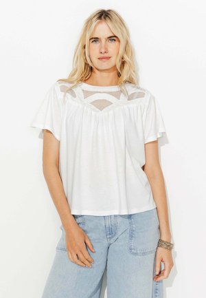 Femme blonde portant un chemisier blanc à manches courtes avec des détails en maille et un jean taille haute bleu clair, debout devant un fond blanc.