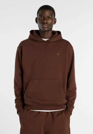 Junger Mann, der einen braunen Hoodie und passende braune Jogginghosen trägt, steht mit den Händen in den Taschen vor einem schlichten weißen Hintergrund.