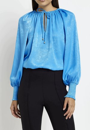 Frau trägt glänzende blaue Bluse mit langen Ärmeln, gerafftem Ausschnitt und Manschetten, kombiniert mit hoch taillierter schwarzer Hose, Frontansicht.