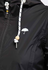 Giacca nera con logo ombrello bianco sul petto, con coulisse bianche con motivi neri, perline in legno e punte in metallo.