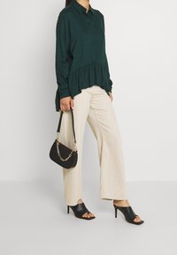 Kvinna som bär en mörkgrön blus med knäppning, beige vida byxor, svarta öppna klackmules, och håller en svart handväska med en guldkedja.