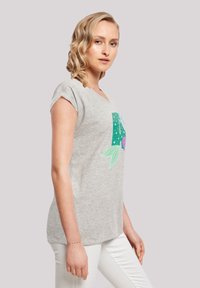 Tricou gri cu mânecă scurtă, având un design floral colorat cu o frunză verde și accente purpurii, purtat cu pantaloni albi.