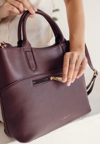 Dunkelviolette Lederhandtasche mit zwei Griffen, einer vorderen Reißverschlusstasche und goldenen Hardware-Akzenten. Glatte Oberfläche mit dezenten Steppnähten.