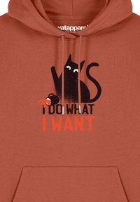 Hoodie couleur rouille avec un graphic de chat noir aux yeux blancs. Le texte indique « JE FAIS CE QUE JE VEUX » en gras orange et noir.