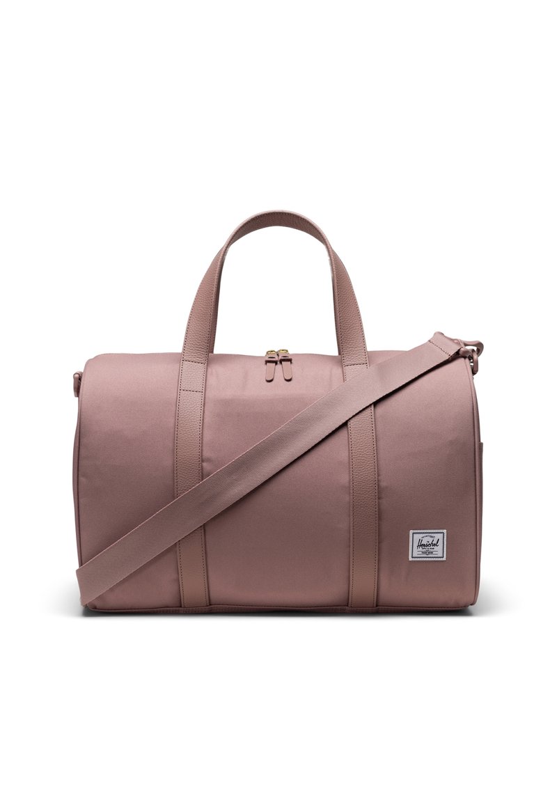 Sac de sport en tissu rose avec des poignées supérieures et une bandoulière amovible. Dispose d'un patch logo à l'avant et d'une surface lisse, légèrement texturée.