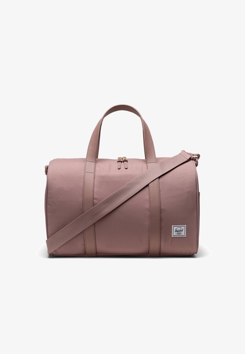 Bolsa de viaje de tela rosa con asas superiores y una correa de hombro desmontable. Presenta un parche con el logo en la parte frontal y una superficie lisa y ligeramente texturizada.