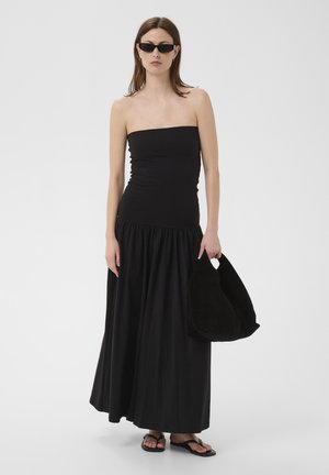 Kobieta w czarnej sukience maxi bez ramiączek, czarnych sandałach, okularach przeciwsłonecznych, trzymająca dużą miękką czarną torebkę, stojąca na jednolitym tle.