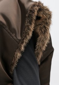 Καφέ εξωτερικό ύφασμα με απαλό επένδυση από συνθετική γούνα στον γιακά· διαθέτει αντίθετη υφή σε μαύρο τμήμα στο μανίκι.