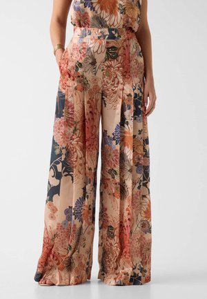 Szerokie spodnie o kwiatowym wzorze w odcieniach brzoskwini, pomarańczy, granatu i fioletu, noszone z jedną ręką w kieszeni i widocznymi gołymi stopami.