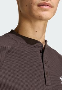 Hnedá košeľa henley s textúrou vafle, okrúhlym golierom, trojitzovou zapínaním a krátkymi raglánovými rukávmi. Obsahuje jemný akcent s logom.