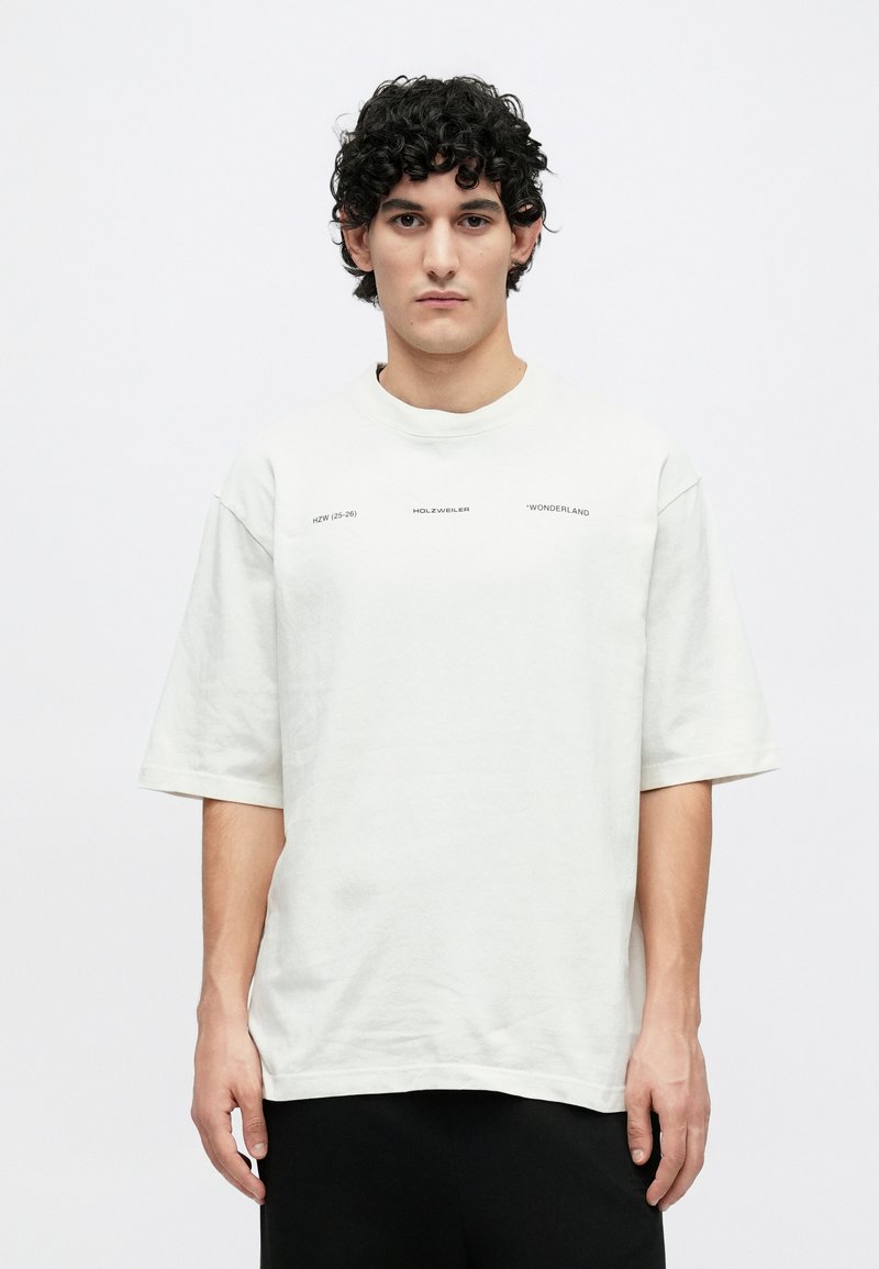 Maglietta oversize bianca in cotone con maniche corte e scollo rotondo. Presenta un testo stampato in nero sul davanti. Texture liscia.