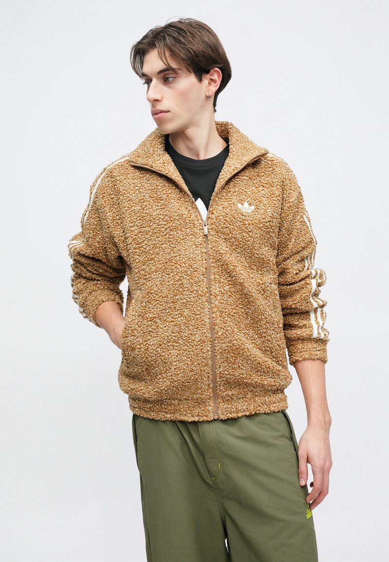 Fuzzy brown zip-up jacket with a textured finish, featuring contrasting white stripes along the sleeves and a small logo on the chest.