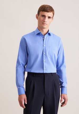 Jeune homme aux cheveux bruns courts portant une chemise bleu clair boutonnée et un pantalon sombre plissé, debout devant un fond clair uni.
