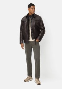 Bruine leren jas met een gewatteerde binnenlaag, crèmekleurig shirt, groene spijkerbroek en beige sneakers met donkerdere accenten, staande pose.