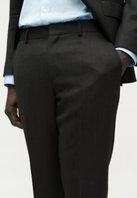 Άνδρας που φοράει σκούρο, ραμμένο κοστούμι παντελόνι και ταιριαστό σακάκι με ανοιχτό γαλάζιο πουκάμισο, με το ένα χέρι στην τσέπη, στέκεται μπροστά σε λευκό φόντο.