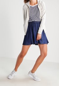 Sudadera gris con cremallera sobre una falda de mezclilla azul marino. La falda tiene un dobladillo desflecado, combinada con zapatillas blancas. Lleva una camiseta de rayas debajo.