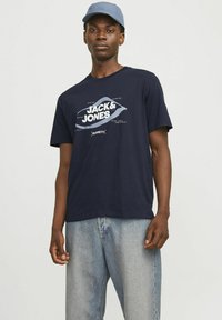 Navyblauwe katoenen T-shirt met grote witte tekst die "JACK & JONES" zegt, gecombineerd met lichtgewassen denim jeans en een blauwe cap.