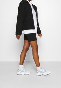 Persoon in zwarte biker shorts, witte hoodie, zwarte gestructureerde jas, witte sokken en wit met lichtblauwe sneakers loopt naar rechts.