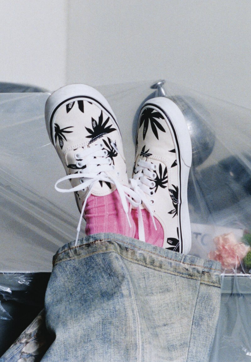 Baskets en toile blanches avec des motifs de feuilles noires, chaussettes côtelées roses et jean bleu, pieds posés sur une surface recouverte de plastique.