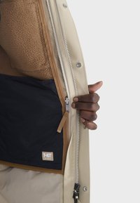 Beige Außenjacke mit Reißverschluss und Druckknöpfen, die ein braunes Fleece-Futter und ein navy Innenfutter zeigt. T-KIT-Label sichtbar.