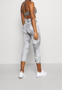 Grijze tie-dye capri legging met hoge tailleband, gecombineerd met een grijze sport-bralette. Model draagt witte sneakers en staat met de rug naar de camera toe.