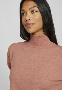 Jasnoróżowy sweter z golfem, wykonany z miękkiej dzianiny, z prążkowanym kołnierzem, luźnym krojem i subtelną teksturą bez wyraźnych wzorów.