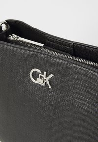 Borsa a tracolla nera con texture, dotata di cerniera argentata e un logo ben in vista. Il materiale sembra sintetico con un motivo a righe.