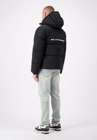 Zwarte pufferjas met capuchon, voorzien van gestikte panelen en witte tekst "BLCK BNDS" op de achterkant. Gecombineerd met lichtblauwe jeans en met een patroon versierde schoenen.