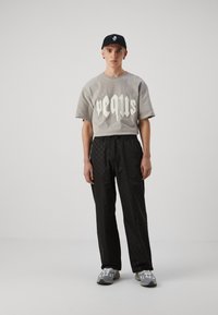 Graues T-Shirt mit kurzen Ärmeln und großem cremefarbenem Grafikdesign, kombiniert mit schwarzen strukturierten Hosen mit einem sechseckigen Muster und grauen Sneakers.