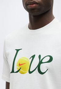 Άνδρας που φοράει λευκό πουκάμισο με τη λέξη «Love» σε πράσινο χρώμα, αντικαθιστώντας το γράμμα «o» με μια κίτρινη μπάλα του τένις που φέρει το κόκκινο λογότυπο Nike.